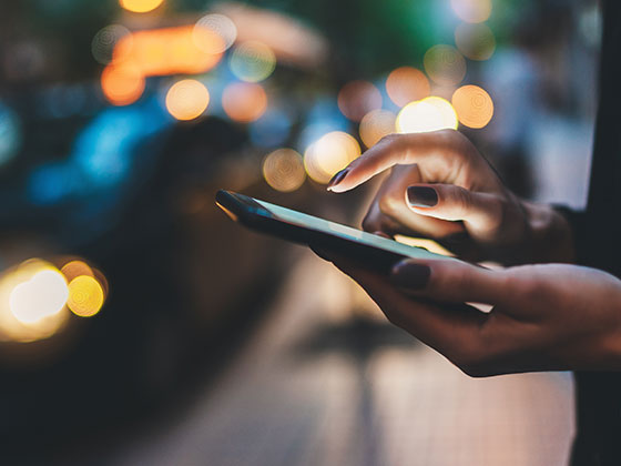 Close up of women's hands using her smart phone while standing on a busy sidewalk in the evening.