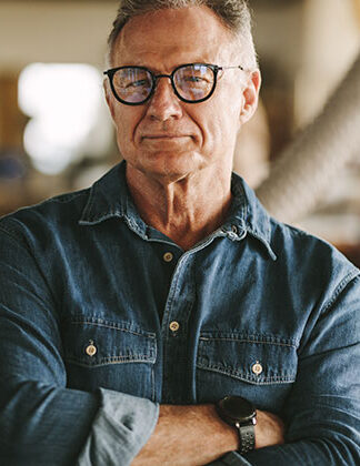 In a woodworking studio, a man smiles at the camera wearing a button up jean shirt with arms crossed. Stock photos of small business owner used to promote winnipeg web designer Sabourin Web & Media.